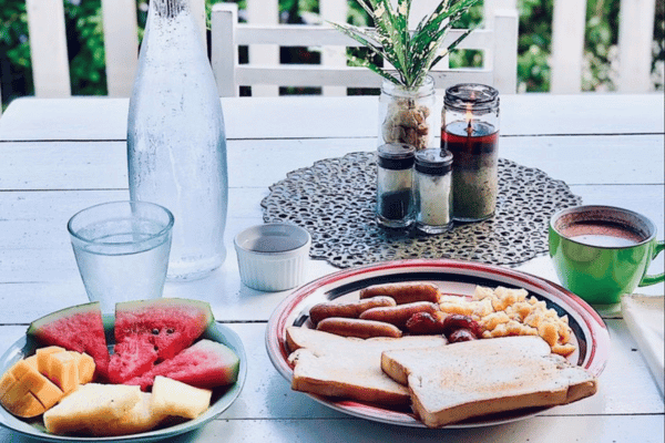 on one plate are toasted white bread and some hotdogs, on another plate are mangoes and watermelon, with a small cup of hot chocolate, and a glass and a bottle of water
