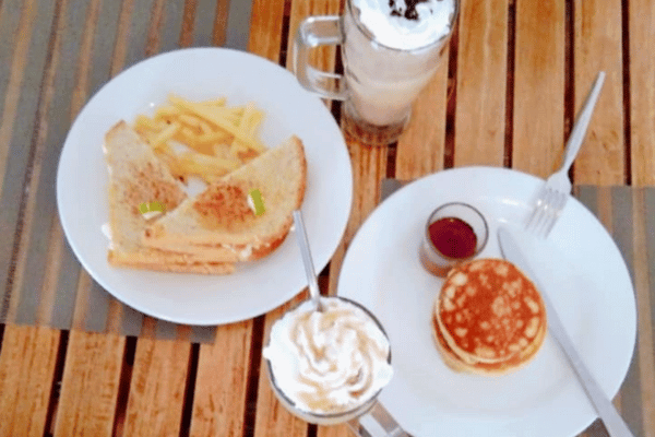 on one plate are some pancakes and syrup, on another plate are some toasted sandwich with french fries. there's also a mug of milk shake and a bottle of whip cream