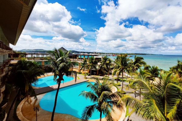 2 swimming pools surrounded by palm trees with the view of the ocean under the blue sky with some white clouds