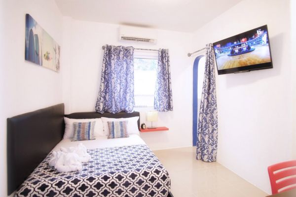 a hotel room with a double size bed with matching bed sheets and curtains, a flat screen tv hanging on the right side wall, an open window with an airconditioning unit above. walls are all white