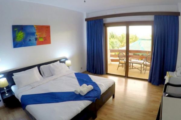 a hotel room with a queen size bed in white sheet with a blue accent matching the blue colored curtains. white walls with a small portrait of a painting at the top of the headboard. a glass door showing the veranda with a table and some chairs