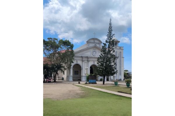 church with spanish colonial design with a watchtower on the right side. the church is artistically hiding behind 2 trees under the blue sky with some white clouds. the ground has patches of green grass